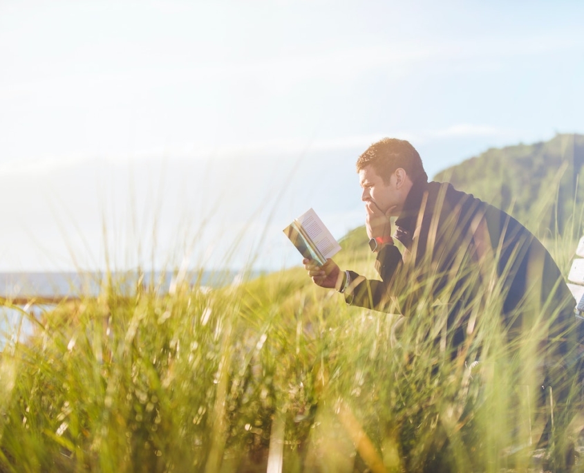 Man sitting on a bench reading a book surrounded by grass with a water view. Reclaim Rehabilitation specializing in reactivation and goal attainment programs to help reclaim function